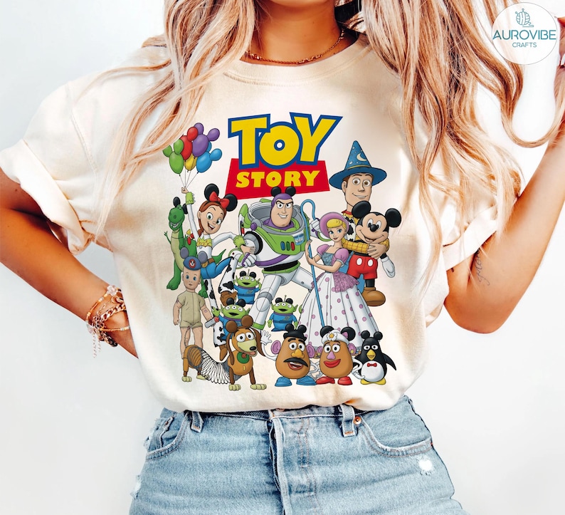 Op de afbeelding: Cr&egrave;mekleurig T-shirt met een kleurrijke afbeelding van personages uit de animatiefilm "Toy Story". Het ontwerp bevat de titel van de film in vette letters en verschillende personages zoals Buzz Lightyear en Woody.