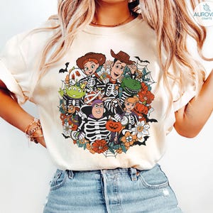 May include: A cream-colored t-shirt featuring a graphic print of animated characters dressed in skeleton costumes. The design includes pumpkins, flowers, spiderwebs, and bats, surrounding the characters in a circular arrangement on the front.