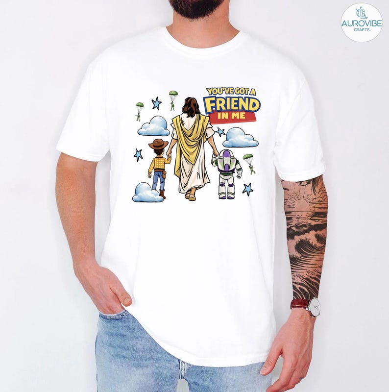 May include: White t-shirt featuring a graphic with the text "YOU'VE GOT A FRIEND IN ME". The graphic depicts a figure in a white robe, holding hands with a cartoon cowboy and a space ranger. Blue clouds, stars, and parachutes are also included in the design.