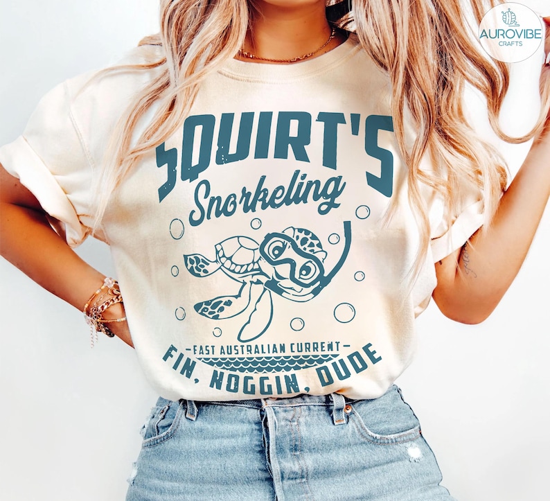 May include: Cream-colored t-shirt with a teal graphic featuring a cartoon turtle wearing a snorkel mask, the words "Squirt's Snorkeling", and the phrase "Fin. Noggin. Dude." The shirt is made of soft material.