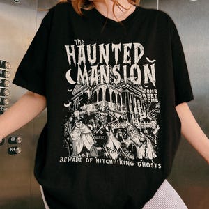 May include: Black t-shirt featuring a graphic of "The Haunted Mansion" with a spooky house, ghosts, and the text "Beware of Hitchhiking Ghosts." The design is in white, creating a contrast against the dark fabric.