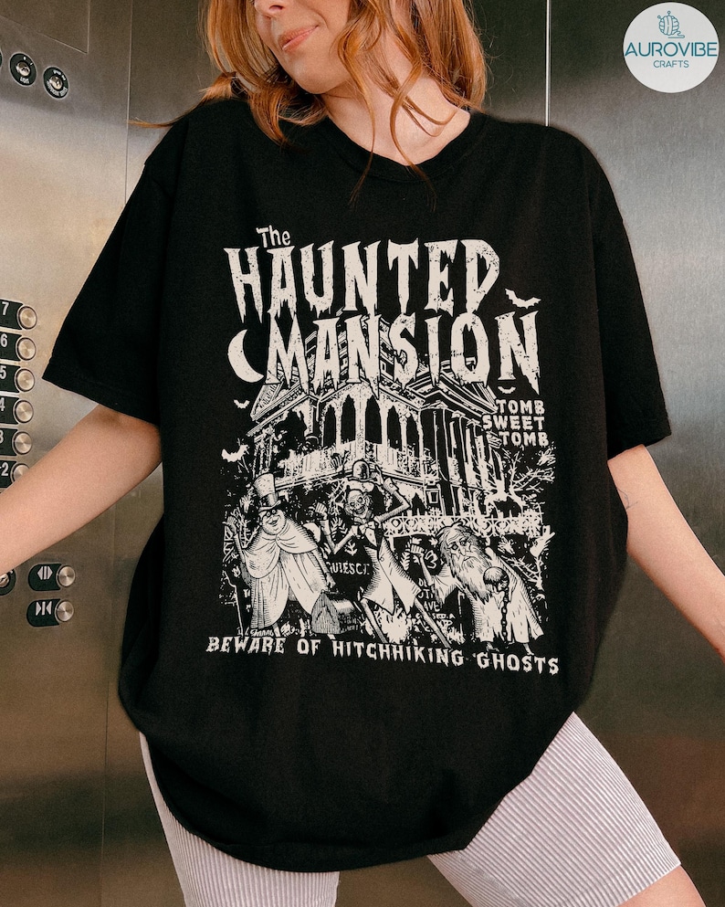 May include: Black t-shirt featuring a graphic of "The Haunted Mansion" with a spooky house, ghosts, and the text "Beware of Hitchhiking Ghosts." The design is in white, creating a contrast against the dark fabric.
