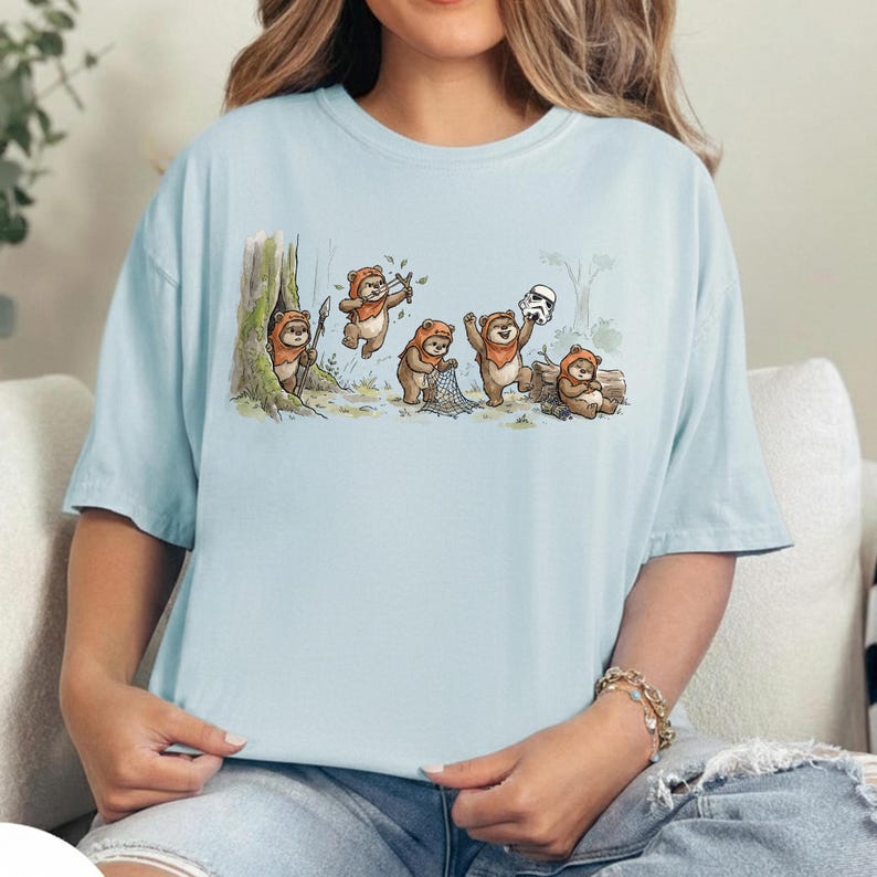 May include: Light blue t-shirt featuring a cartoon illustration of Ewoks in a forest setting. The design shows several Ewoks interacting with a Stormtrooper helmet and a net. The Ewoks are depicted in their traditional brown fur and hooded attire.