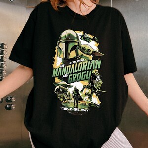 Pode incluir: Camiseta preta com um gr&aacute;fico de Star Wars com "The Mandalorian and Grogu". O design inclui o capacete do Mandaloriano, Grogu e a frase "This is the Way".