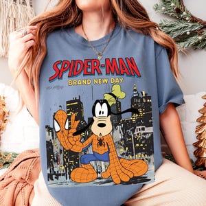 May include: A blue t-shirt featuring a graphic of Spider-Man and Goofy in a city setting. The text "SPIDER-MAN" is in red with a blue outline, and "BRAND NEW DAY" is below it. Goofy is dressed as Spider-Man.