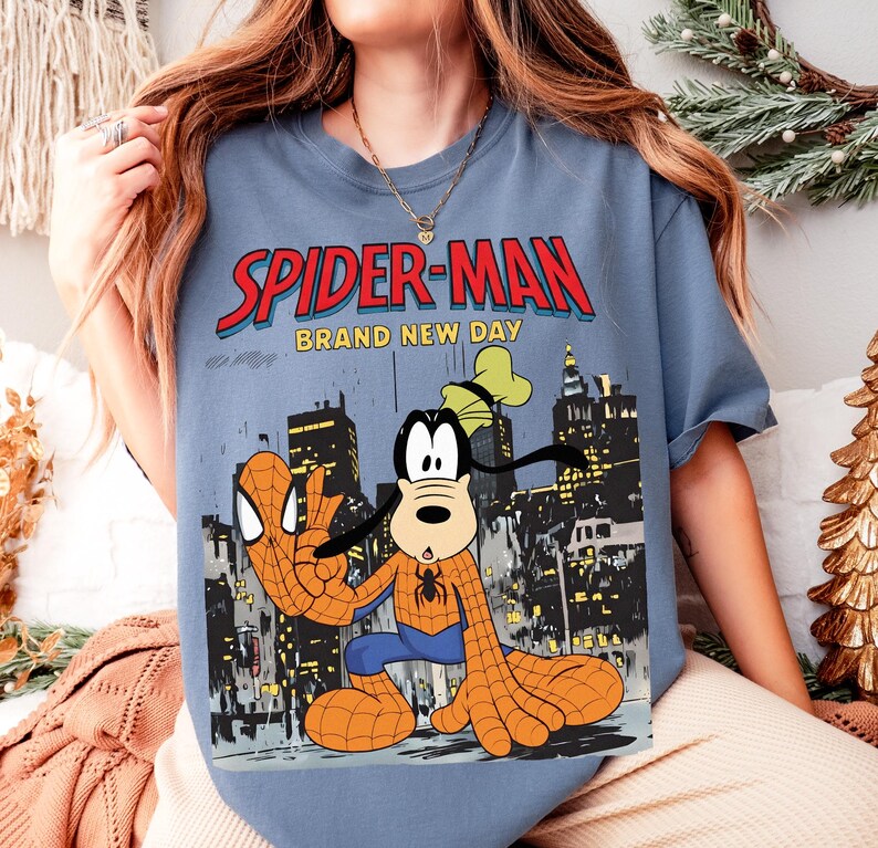 May include: A blue t-shirt featuring a graphic of Spider-Man and Goofy in a city setting. The text "SPIDER-MAN" is in red with a blue outline, and "BRAND NEW DAY" is below it. Goofy is dressed as Spider-Man.