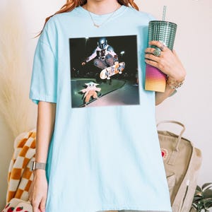 Puede incluir: Camiseta azul claro con un estampado gr&aacute;fico de un skater y un personaje peque&ntilde;o. El gr&aacute;fico est&aacute; sobre un fondo oscuro. La camiseta es de color azul claro.