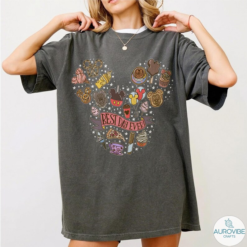 May include: A charcoal gray short-sleeve t-shirt featuring a graphic print of various snacks and treats arranged in a Mickey Mouse head shape. A pink banner across the center contains the text "BEST DAY EVER!" surrounded by small white stars.