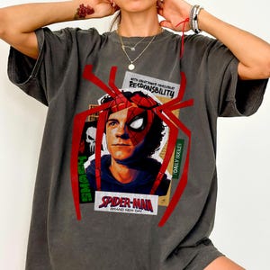 Op de afbeelding: Donkergrijs t-shirt met een Spider-Man-afbeelding. Het ontwerp bevat een portret van Spider-Man, een rood spinnenweb en de tekst "SPIDER-MAN BRAND NEW DAY" en "WITH GREAT POWER COMES GREAT RESPONSIBILITY".