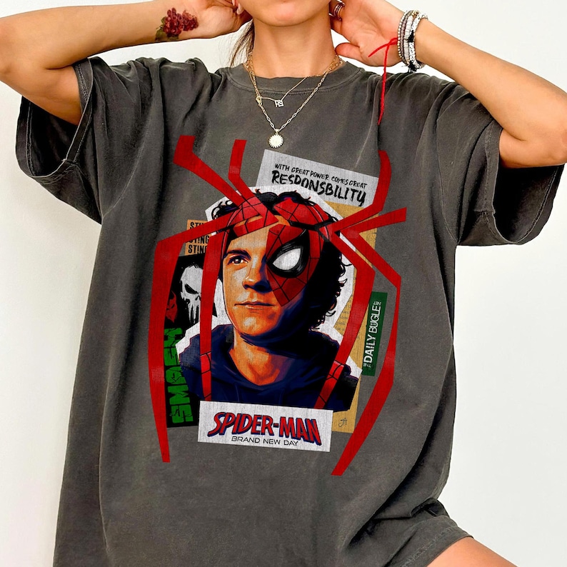 Op de afbeelding: Donkergrijs t-shirt met een Spider-Man-afbeelding. Het ontwerp bevat een portret van Spider-Man, een rood spinnenweb en de tekst "SPIDER-MAN BRAND NEW DAY" en "WITH GREAT POWER COMES GREAT RESPONSIBILITY".