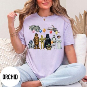 Peut inclure: Un t-shirt violet clair avec des personnages et des v&eacute;hicules de Star Wars, dont R2-D2, Chewbacca, C-3PO, Dark Vador, un Stormtrooper et Baby Yoda. Le t-shirt porte &eacute;galement le texte "ORCHID" en bas.