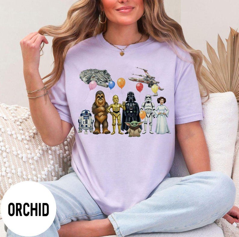 Peut inclure: Un t-shirt violet clair avec des personnages et des v&eacute;hicules de Star Wars, dont R2-D2, Chewbacca, C-3PO, Dark Vador, un Stormtrooper et Baby Yoda. Le t-shirt porte &eacute;galement le texte "ORCHID" en bas.