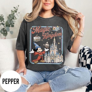 May include: Dark gray t-shirt with a graphic featuring Mickey Mouse in a wizard outfit, the Magic Kingdom logo, a castle, and other park icons. The shirt is a relaxed fit.
