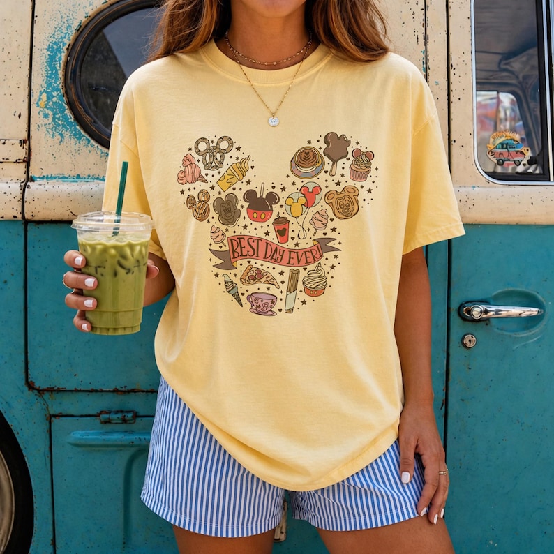 May include: A pale yellow short-sleeve t-shirt featuring a graphic of various snacks and treats arranged in a Mickey Mouse head shape, with a banner across the center that reads BEST DAY EVER in red letters, paired with blue and white striped shorts.