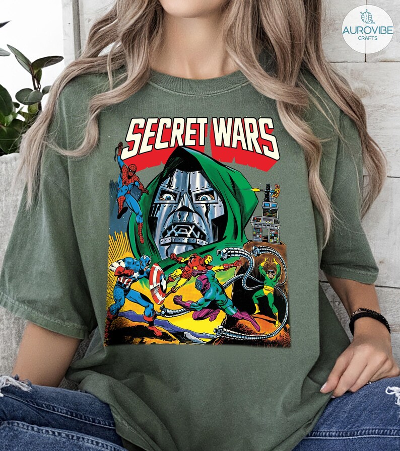 May include: Olive green t-shirt featuring a comic book graphic. The graphic includes the text "SECRET WARS" in red, and illustrations of various superheroes and villains. The design is reminiscent of vintage comic book art.