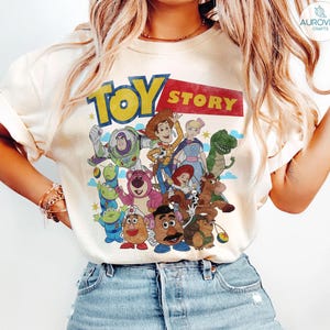 May include: Cream-colored t-shirt featuring the "Toy Story" logo in yellow and red, with a group of cartoon characters. The design includes Buzz Lightyear, Woody, and other characters from the animated film. The shirt is a casual wear item.