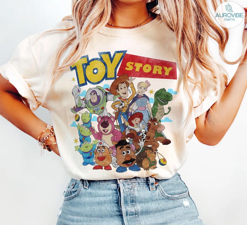 May include: Cream-colored t-shirt featuring the "Toy Story" logo in yellow and red, with a group of cartoon characters. The design includes Buzz Lightyear, Woody, and other characters from the animated film. The shirt is a casual wear item.
