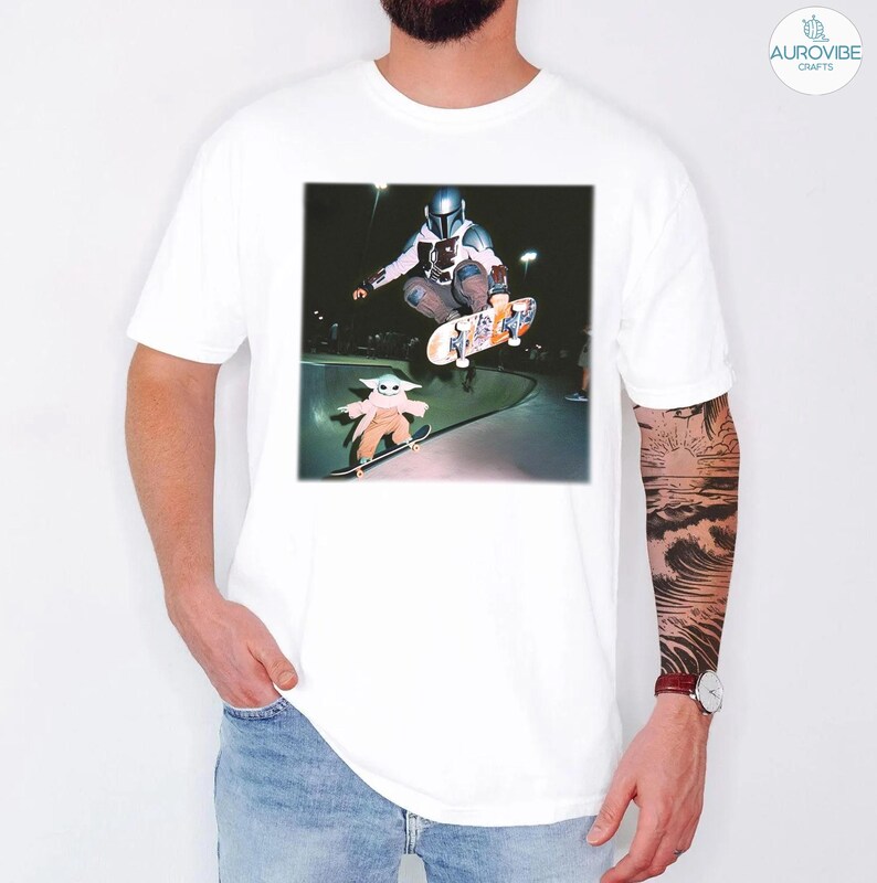 Puede incluir: Camiseta blanca con un gr&aacute;fico de un skater con casco y armadura en el aire, con una figura m&aacute;s peque&ntilde;a de un personaje en monopat&iacute;n debajo. La imagen est&aacute; ambientada en un skate park. El logotipo de Aurovibe Crafts est&aacute; en la esquina superior derecha.