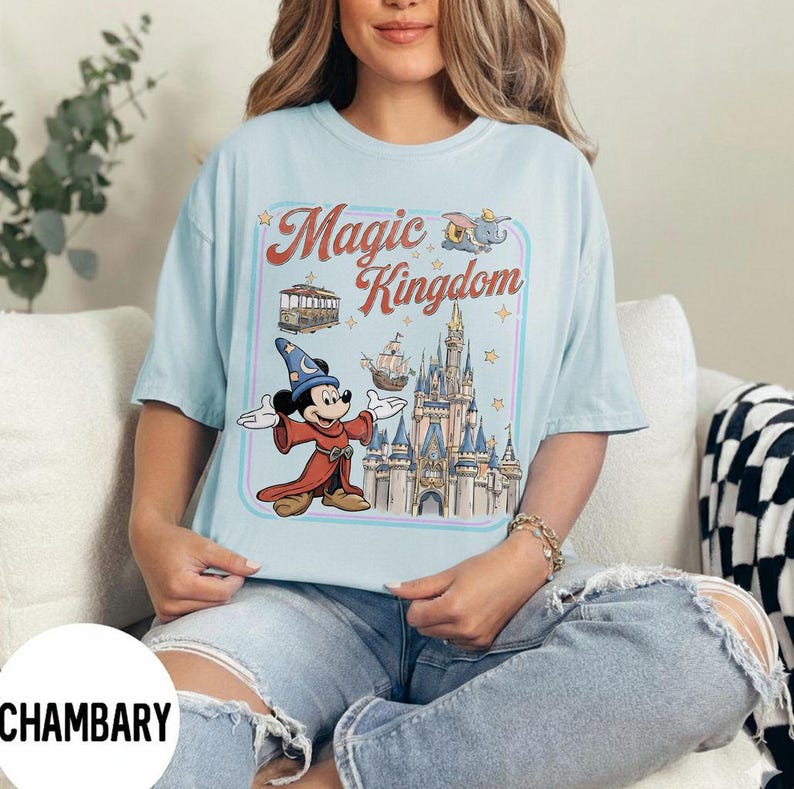 May include: Light blue t-shirt featuring a graphic of Mickey Mouse in a sorcerer's outfit, with the words "Magic Kingdom" in a retro font. The design includes a castle, a trolley, and a ship. The shirt is a comfortable, casual style.