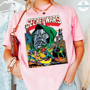 May include: A light pink t-shirt featuring a comic book graphic with the words "SECRET WARS" in red and white. The graphic depicts various superheroes and villains in action, with a metallic-faced character prominently displayed.