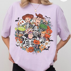 May include: A light purple short-sleeve t-shirt featuring a graphic print of various cartoon characters dressed in skeleton costumes. The design includes pumpkins, flowers, spiderwebs, and bats, with the characters arranged in a circular cluster.