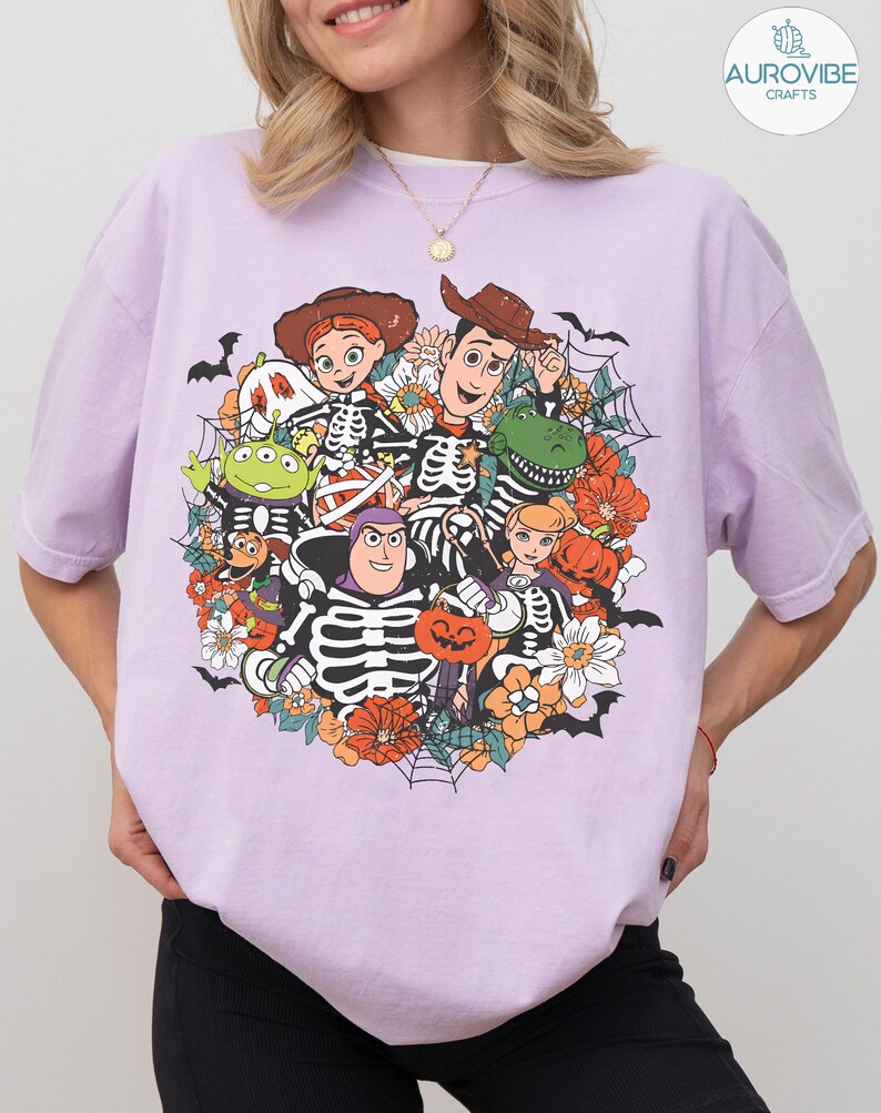 May include: A light purple short-sleeve t-shirt featuring a graphic print of various cartoon characters dressed in skeleton costumes. The design includes pumpkins, flowers, spiderwebs, and bats, with the characters arranged in a circular cluster.