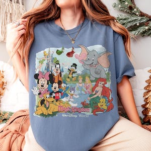 Puede incluir: Una camiseta azul con un gr&aacute;fico colorido de personajes de Disney, incluyendo Mickey Mouse, Minnie Mouse, Dumbo y Ariel. El texto "Magic Kingdom Walt Disney World" est&aacute; impreso debajo de los personajes.
