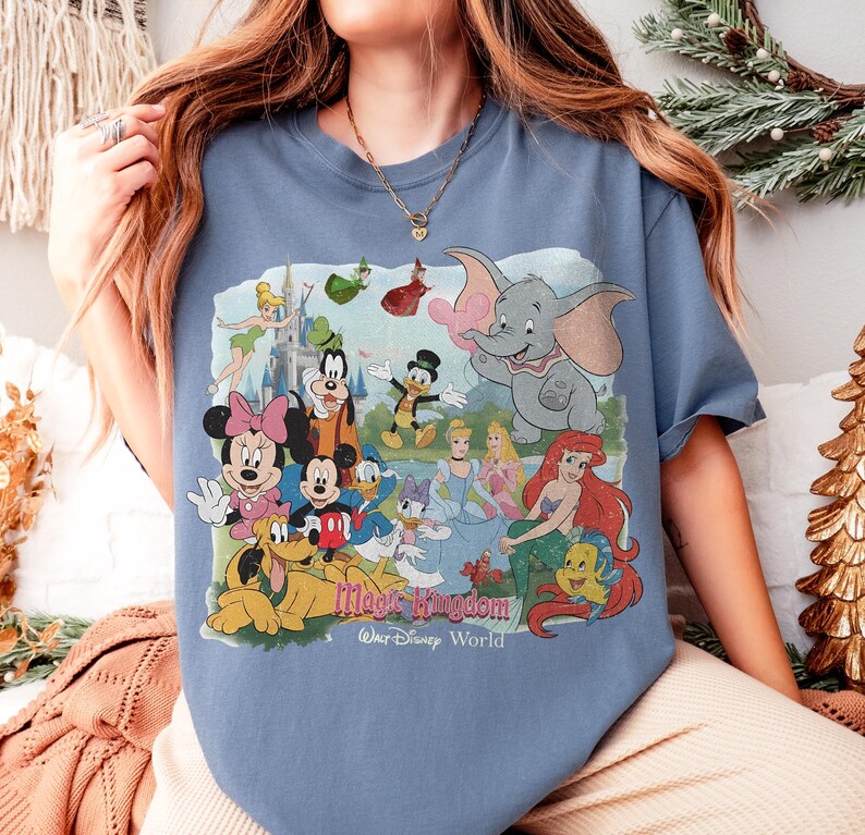 Puede incluir: Una camiseta azul con un gr&aacute;fico colorido de personajes de Disney, incluyendo Mickey Mouse, Minnie Mouse, Dumbo y Ariel. El texto "Magic Kingdom Walt Disney World" est&aacute; impreso debajo de los personajes.