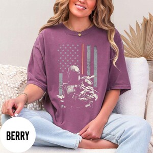 May include: Berry-colored t-shirt featuring a distressed American flag design with a character illustration. The design includes stars, stripes, and a character silhouette. The shirt is a short-sleeved, crew neck style.