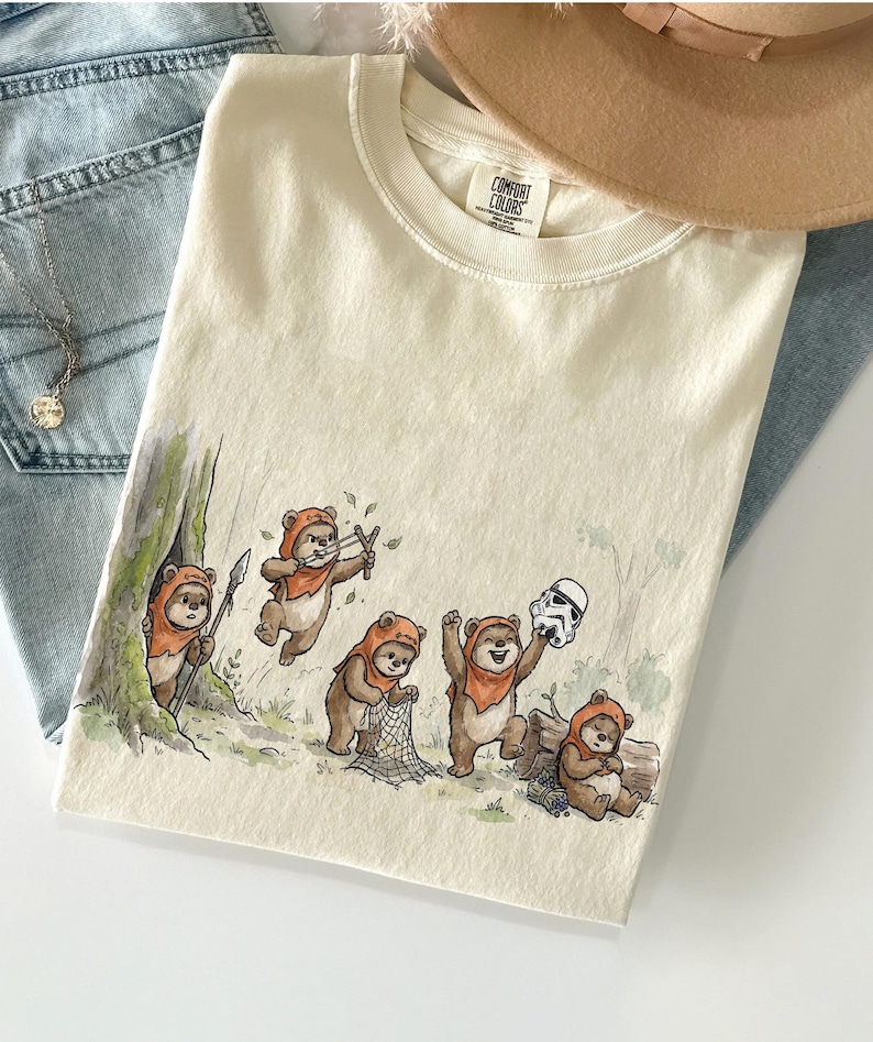 May include: Cream-colored t-shirt featuring a whimsical illustration of Ewoks in a forest setting. The design includes Ewoks with slingshots, nets, and a Stormtrooper helmet. The shirt has a Comfort Colors tag.