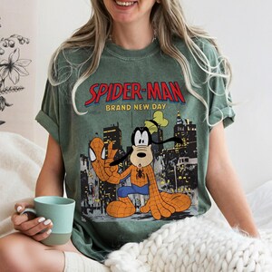 May include: Green t-shirt featuring a graphic of Spider-Man and Goofy in a city setting. The text "SPIDER-MAN BRAND NEW DAY" is printed across the top. The shirt has rolled-up sleeves and a relaxed fit.