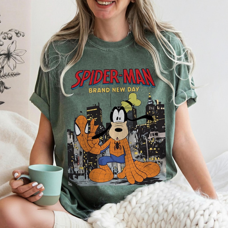 May include: Green t-shirt featuring a graphic of Spider-Man and Goofy in a city setting. The text "SPIDER-MAN BRAND NEW DAY" is printed across the top. The shirt has rolled-up sleeves and a relaxed fit.