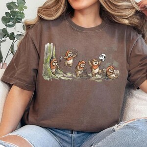 May include: Brown t-shirt featuring a cartoon illustration of Ewoks in a forest setting. The Ewoks are depicted in various poses, including holding spears, nets, and a Stormtrooper helmet. The shirt has a vintage, worn look.