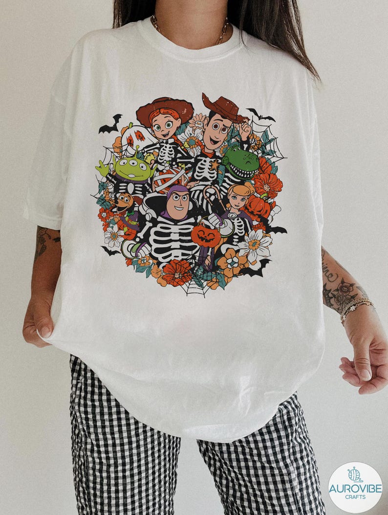 May include: A white t-shirt featuring a colorful graphic print of various animated characters dressed in skeleton costumes. The design includes orange flowers, spider webs, bats, and pumpkins, creating a festive Halloween-themed illustration.