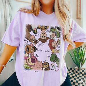 K&ouml;nnte beinhalten: Ein helllila T-Shirt mit einem farbenfrohen Aufdruck verschiedener Charaktere aus dem Film Toy Story. Die Grafik zeigt Woody, Buzz Lightyear, Jessie und andere, mit ihren Namen darunter. Das Shirt besteht aus einem weichen Material.