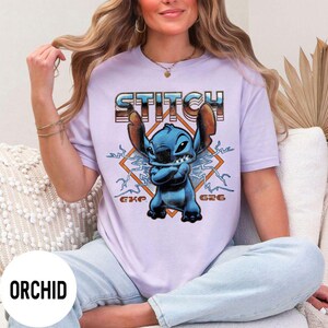 Puede incluir: Una camiseta de color lila claro con un gr&aacute;fico del personaje Stitch de Lilo & Stitch. La palabra "STITCH" est&aacute; impresa encima del personaje en una fuente retro. El personaje es azul con los brazos cruzados, sobre un dise&ntilde;o en forma de diamante.