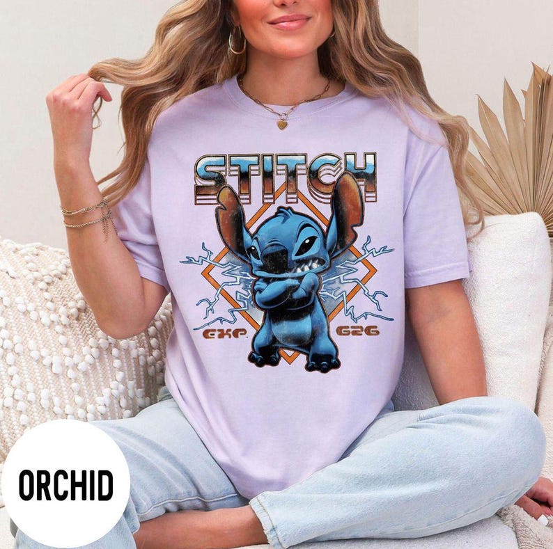 Puede incluir: Una camiseta de color lila claro con un gr&aacute;fico del personaje Stitch de Lilo & Stitch. La palabra "STITCH" est&aacute; impresa encima del personaje en una fuente retro. El personaje es azul con los brazos cruzados, sobre un dise&ntilde;o en forma de diamante.