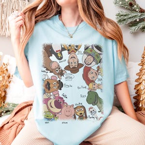 K&ouml;nnte beinhalten: Hellblaues T-Shirt mit einem farbenfrohen Grafikdesign von Charakteren aus dem Animationsfilm Toy Story. Das Design zeigt Woody, Buzz Lightyear, Jessie und andere beliebte Charaktere. Die Namen der Charaktere sind in einer verspielten Schriftart geschrieben.