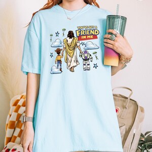 May include: Light blue t-shirt with a graphic featuring a religious figure and cartoon characters. The text "YOU'VE GOT A FRIEND IN ME" is displayed in a yellow and red banner. The design includes stars, clouds, and parachutes.