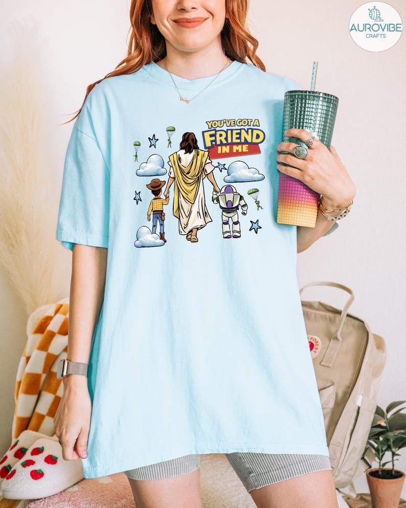 May include: Light blue t-shirt with a graphic featuring a religious figure and cartoon characters. The text "YOU'VE GOT A FRIEND IN ME" is displayed in a yellow and red banner. The design includes stars, clouds, and parachutes.