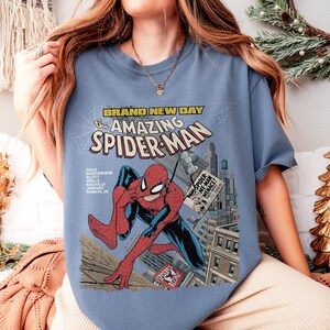 May include: A light blue t-shirt featuring a comic book cover design of Spider-Man. The graphic includes the text "BRAND NEW DAY" and "the AMAZING SPIDER-MAN". The illustration shows Spider-Man in a dynamic pose with a cityscape background.