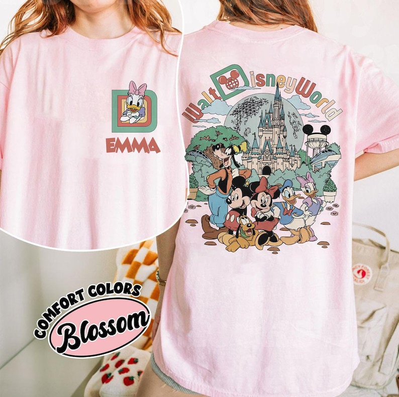 May include: A light pink t-shirt featuring a Daisy Duck graphic with the name "EMMA" on the front. The back of the shirt displays a colorful "Walt Disney World" design with Mickey Mouse and friends. The shirt is part of the "Comfort Colors Blossom" collection.