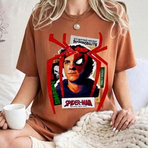 Op de afbeelding: Een roestkleurig t-shirt met een Spider-Man-afbeelding. Het ontwerp bevat een portret van het personage, een spinnenweb en de tekst "SPIDER-MAN BRAND NEW DAY" en "WITH GREAT POWER COMES GREAT RESPONSIBILITY".