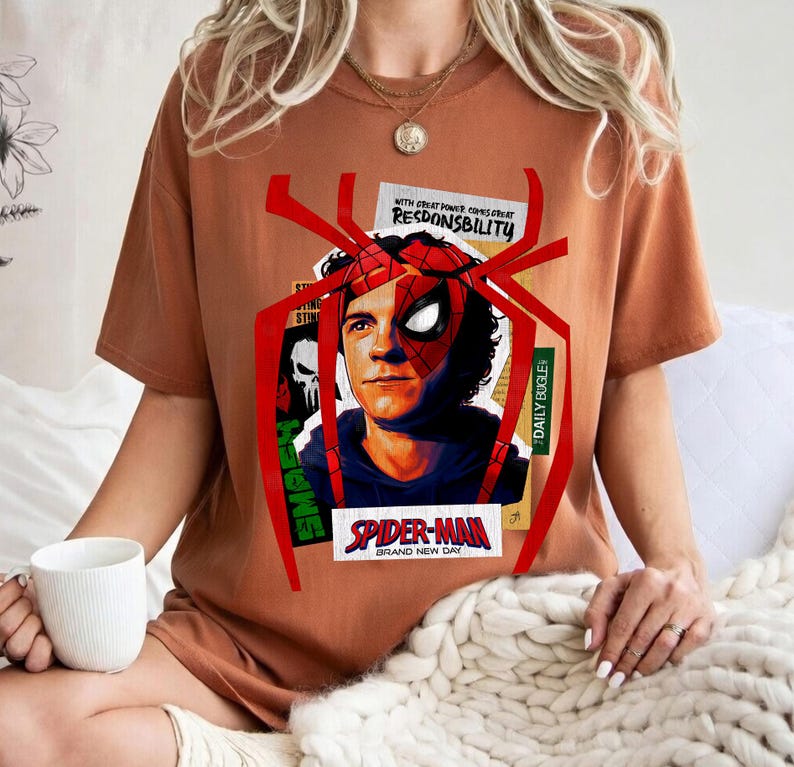 Op de afbeelding: Een roestkleurig t-shirt met een Spider-Man-afbeelding. Het ontwerp bevat een portret van het personage, een spinnenweb en de tekst "SPIDER-MAN BRAND NEW DAY" en "WITH GREAT POWER COMES GREAT RESPONSIBILITY".