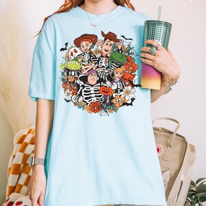 May include: A light blue short-sleeve t-shirt featuring a colorful graphic print of various animated characters dressed in skeleton costumes, surrounded by orange pumpkins, white flowers, and black spiderwebs on a light blue background.