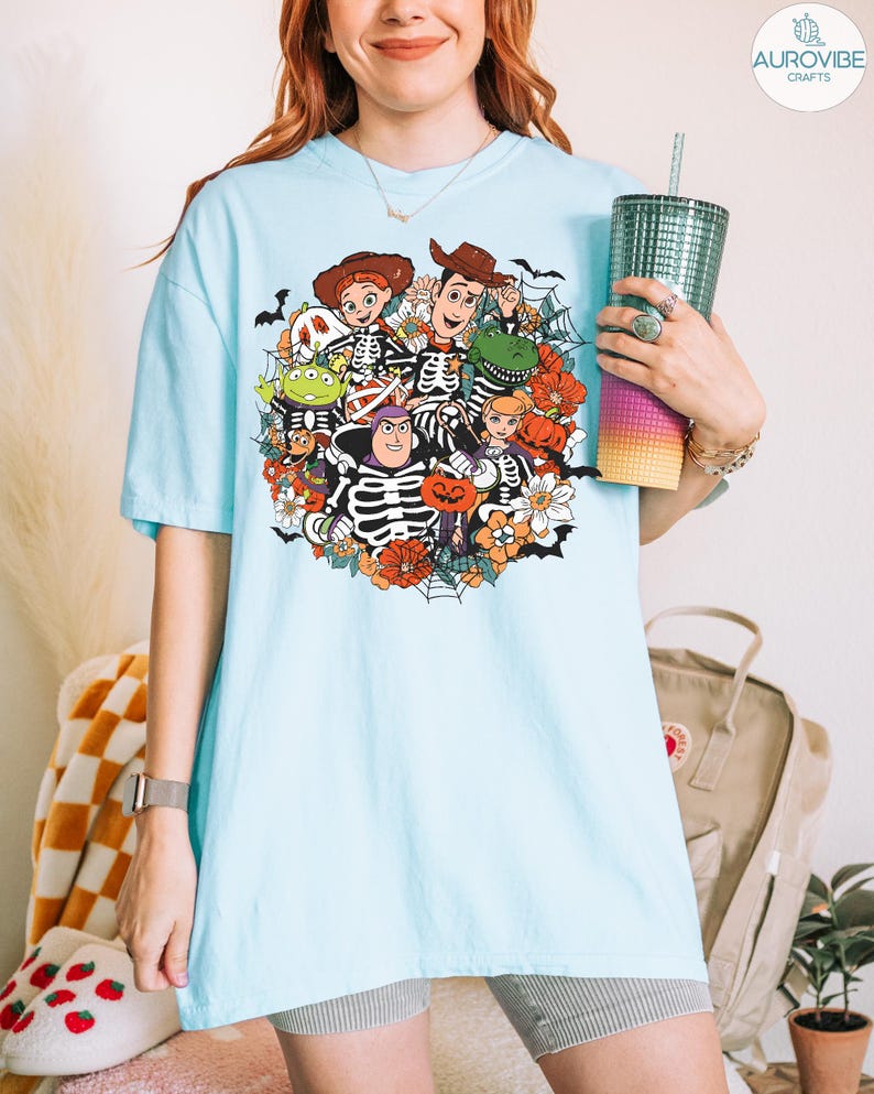 May include: A light blue short-sleeve t-shirt featuring a colorful graphic print of various animated characters dressed in skeleton costumes, surrounded by orange pumpkins, white flowers, and black spiderwebs on a light blue background.