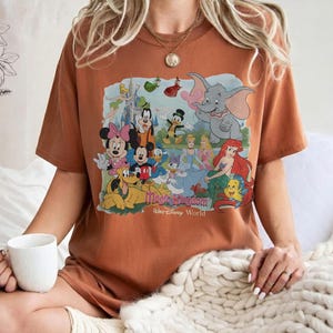 Puede incluir: Camiseta de color &oacute;xido con un gr&aacute;fico colorido de personajes de Disney, incluyendo Mickey Mouse y Dumbo. El dise&ntilde;o incluye el texto "Magic Kingdom" y "Walt Disney World". La camiseta tiene mangas cortas.