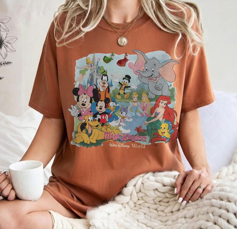 Puede incluir: Camiseta de color &oacute;xido con un gr&aacute;fico colorido de personajes de Disney, incluyendo Mickey Mouse y Dumbo. El dise&ntilde;o incluye el texto "Magic Kingdom" y "Walt Disney World". La camiseta tiene mangas cortas.