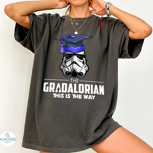 Pu&ograve; includere: T-shirt grigio scuro con un casco da Stormtrooper con cappello di laurea. Il testo bianco recita "THE GRADALORIAN THIS IS THE WAY". La maglietta ha maniche corte e una vestibilit&agrave; ampia.