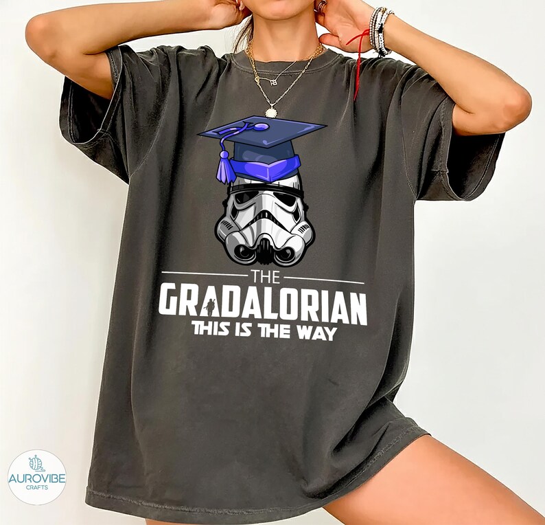 Pu&ograve; includere: T-shirt grigio scuro con un casco da Stormtrooper con cappello di laurea. Il testo bianco recita "THE GRADALORIAN THIS IS THE WAY". La maglietta ha maniche corte e una vestibilit&agrave; ampia.